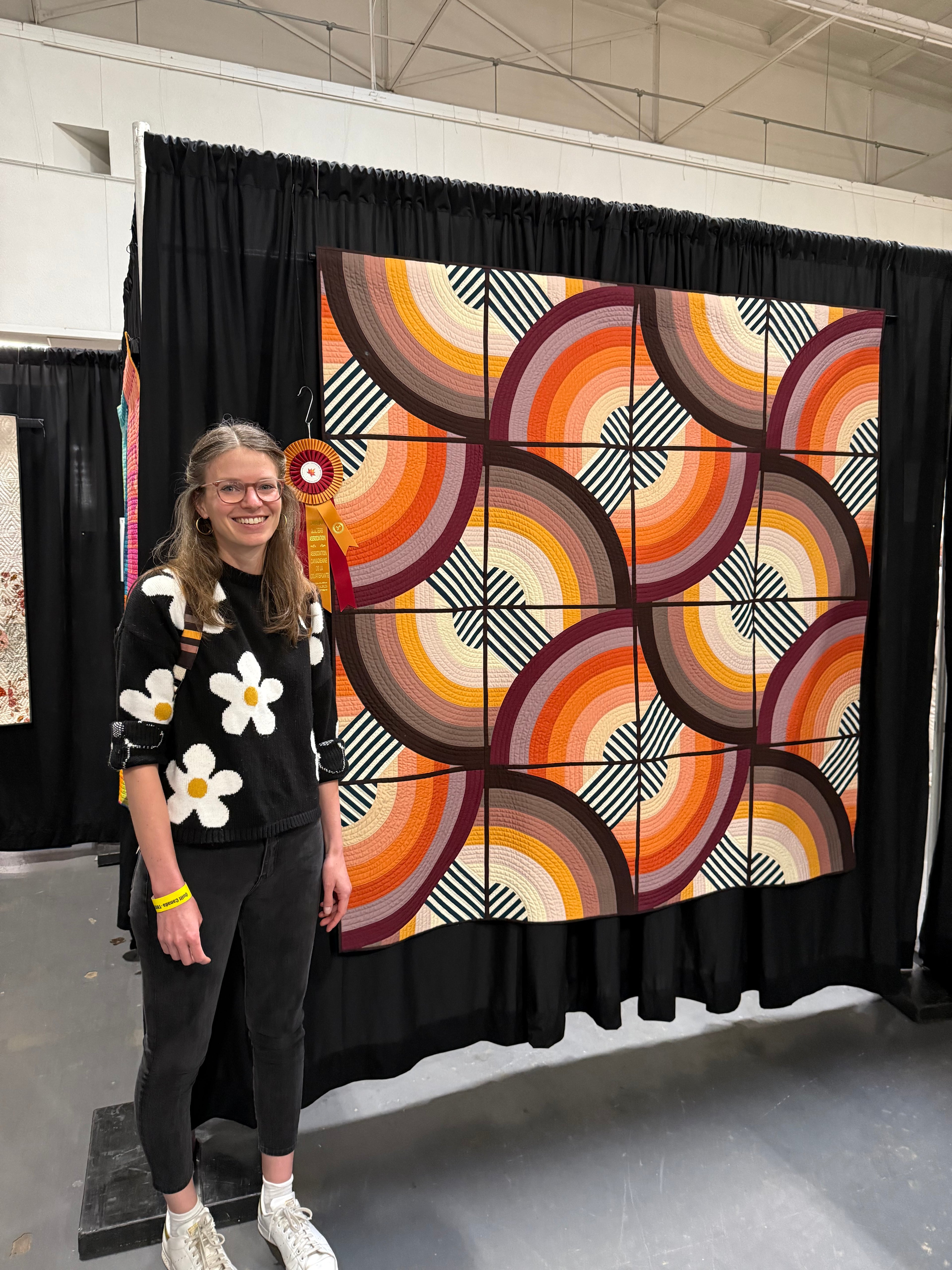 Person standing next to a geometric quilt hanging against a black curtain. A ribbon is hung next to the quilt.