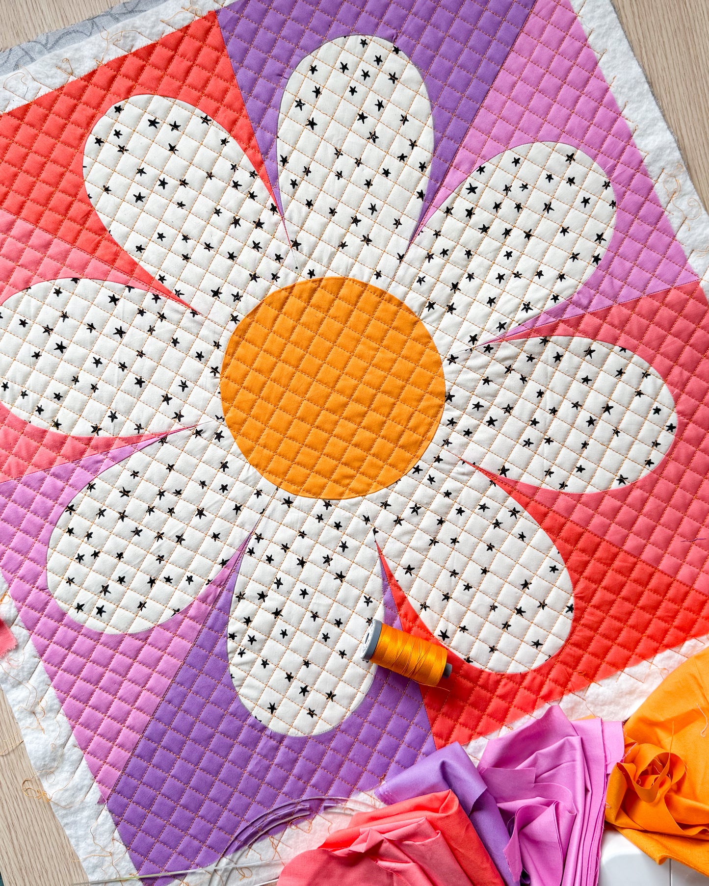 Daisy block made with a yellow center, white fabric with black star petals, and the background is made of purples and pinks.