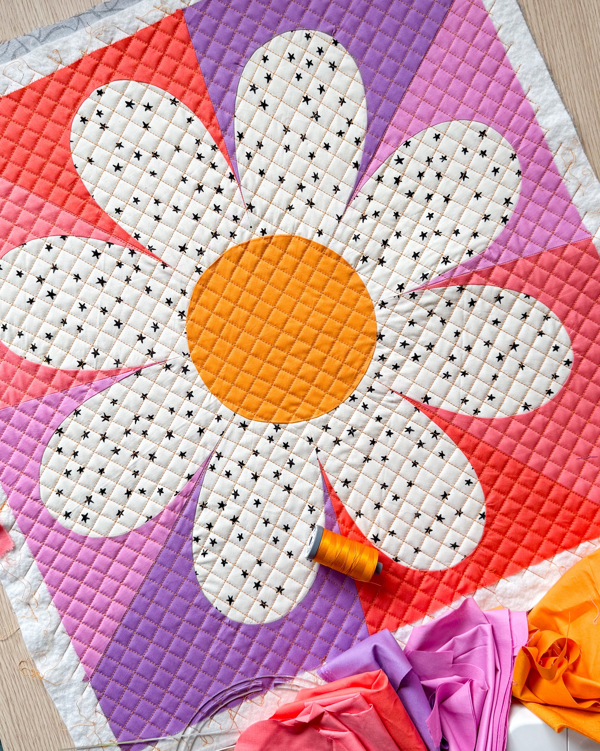 Daisy block made with a yellow center, white fabric with black star petals, and the background is made of purples and pinks.