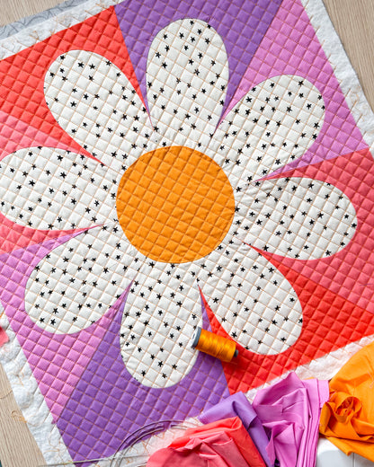 Daisy block made with a yellow center, white fabric with black star petals, and the background is made of purples and pinks.