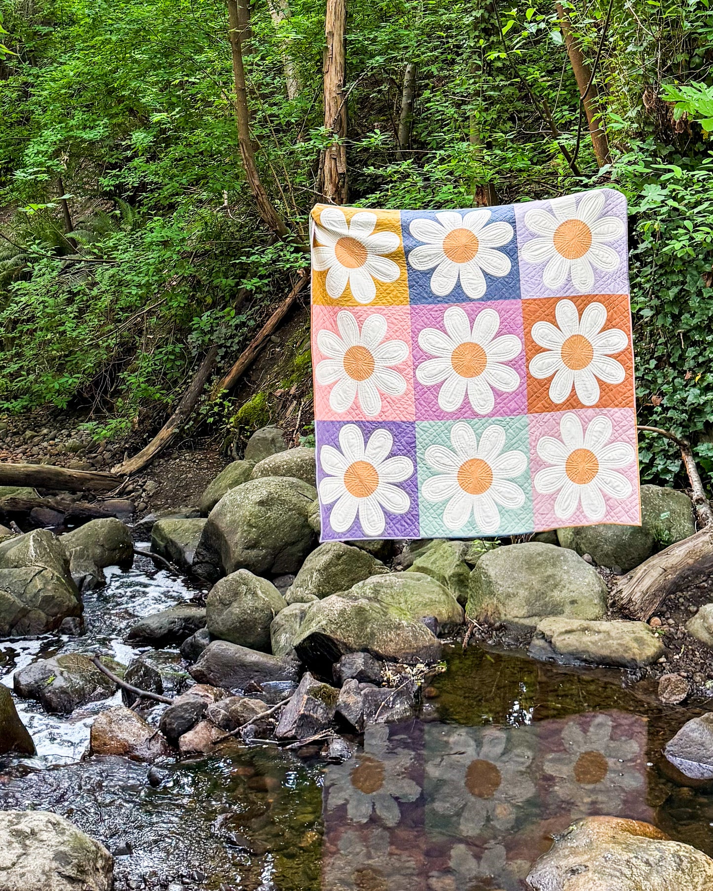 Daisy quilt held up in a forest next to a stream. 