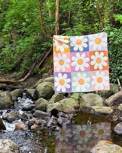 Daisy quilt held up in a forest next to a stream. 