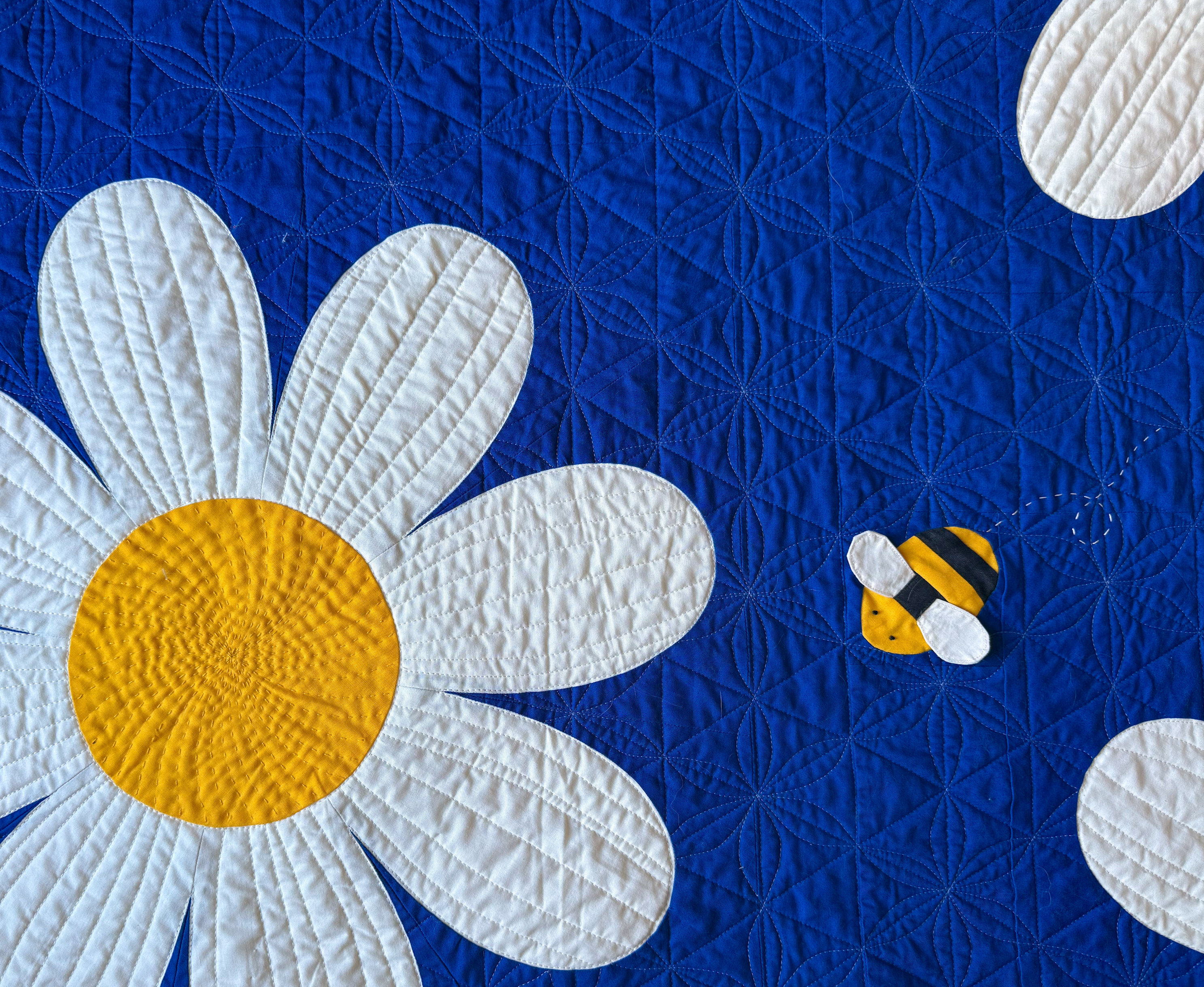 Close up view of a quilt with a large daisy and bee on a blue background.